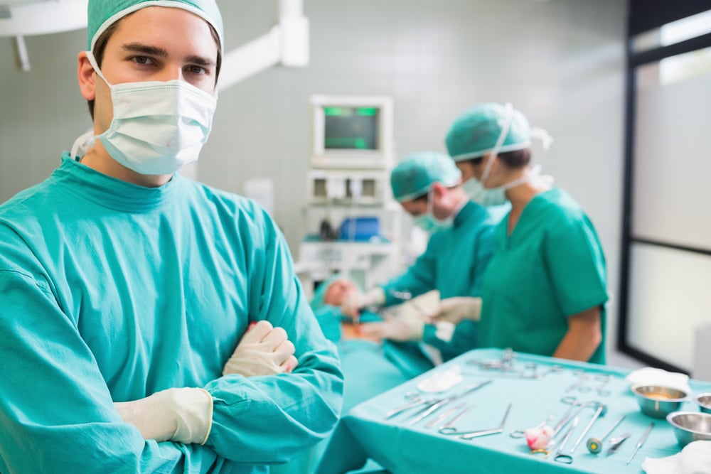 Surgeon with arms crossed looking at camera with colleagues performing in background Surgeon with arms crossed looking at camera with colleagues performing in background